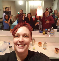 Smiling person takes a selfie at a lively indoor party as a group behind them toasts with teal cocktails over a countertop bar with copper mugs, mixers, and bar tools.
