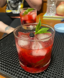 Bright red blackberry-mint cocktail with ice in a lowball glass, garnished with a blackberry and mint leaf, sitting on a black bar mat with a second blurred drink and home table in the background.