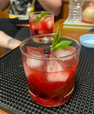 Bright red blackberry-mint cocktail with ice in a lowball glass, garnished with a blackberry and mint leaf, sitting on a black bar mat with a second blurred drink and home table in the background.