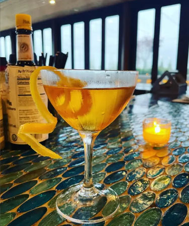 Golden cocktail in a coupe glass with a long lemon twist on a blue-green mosaic bar top, lit votive candle and bottles by large windows in an upscale lounge.