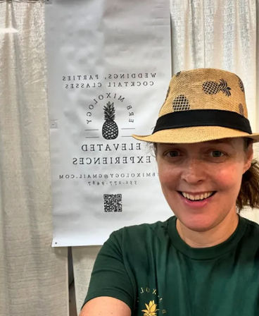 Smiling person in a straw hat with pineapple print and green T‑shirt taking a selfie in front of a white indoor event booth banner featuring a pineapple logo.