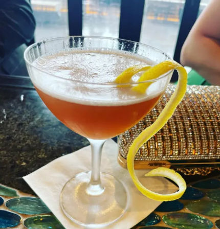 Amber cocktail in a coupe glass with frothy top and long lemon twist garnish, resting on a napkin at an outdoor waterfront bar