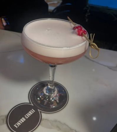 Frothy pink cocktail in a coupe glass topped with creamy foam and a red-berry bamboo pick, served on a marble bar counter in a dimly lit cocktail bar.
