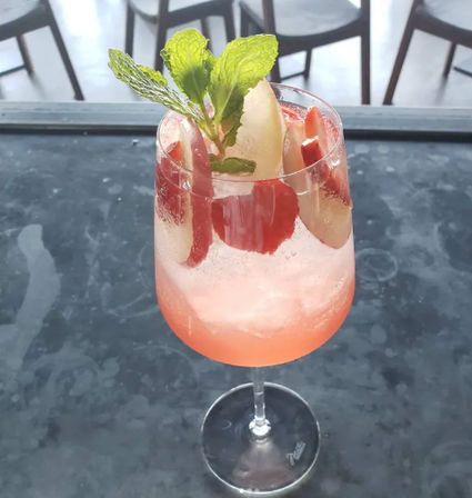 Pink sparkling strawberry cocktail in a stemmed wine glass, garnished with fresh mint and fruit slices, sitting on a dark marble café table — refreshing summer spritz.