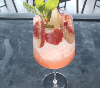 Pink sparkling strawberry cocktail in a stemmed wine glass, garnished with fresh mint and fruit slices, sitting on a dark marble café table — refreshing summer spritz.