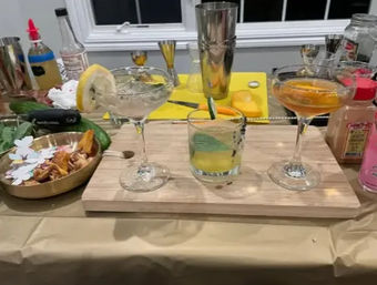 Three craft cocktails on a wooden board at a home bar: two citrus‑garnished coupe glasses and a cucumber‑topped rocks glass, surrounded by a metal shaker, jigger, sliced fruit and bottles.