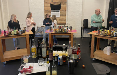 Loft-style mixology workshop in a white-brick studio with wooden prep tables loaded with liquor bottles, mixers, eggs, strawberries and cocktail tools while a small group of adults chat and prepare drinks.