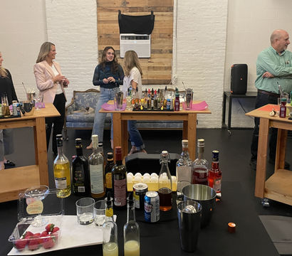 Loft-style mixology workshop in a white-brick studio with wooden prep tables loaded with liquor bottles, mixers, eggs, strawberries and cocktail tools while a small group of adults chat and prepare drinks.