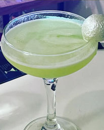 Bright green frothy cocktail in a coupe glass with a sugared rim and a small round garnish on the rim, close-up on a bar counter.