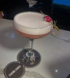 Pink frothy cocktail in a coupe glass topped with creamy foam and a sugared red-berry skewer, served on a marble bar countertop with a round coaster.