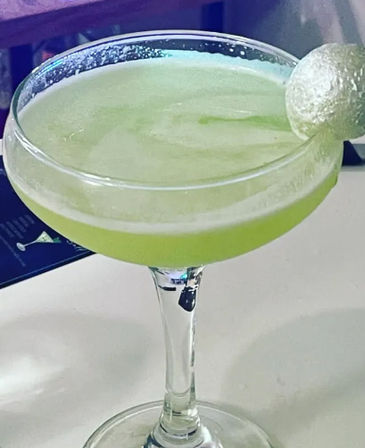 Bright frosty green cocktail in a coupe glass with light foam, garnished with a small lime-like ball on the rim, on a white bar surface.