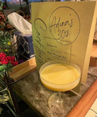 Frothy yellow cocktail in a coupe glass on a marble bar countertop beside a hand-lettered yellow menu marked “30th,” with festive red berries and a tissue box in the background.
