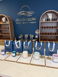 Coastal-themed bath and home boutique interior with a navy wall and gold wave logo, wooden arched shelving filled with amber-jar candles and colorful votives, blue gift bags on the counter and candle sample jars on clipboards.
