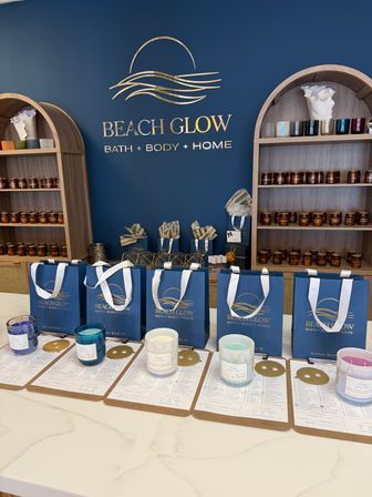 Coastal-themed bath and home boutique interior with a navy wall and gold wave logo, wooden arched shelving filled with amber-jar candles and colorful votives, blue gift bags on the counter and candle sample jars on clipboards.