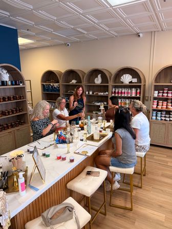 Group of women at a bright boutique wellness studio taking a candle and skincare workshop around a marble-topped island with jars, lotions and wooden arched shelving.