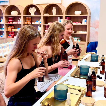 Three women at a bright boutique fragrance bar sampling scented oils from amber bottles during a candle-making workshop with wooden arched shelves of colorful jars