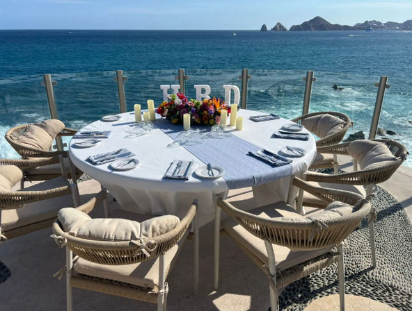 Sunlit oceanfront dining table set for six on a seaside terrace with cushioned wicker chairs, white linen, floral centerpiece and candles overlooking turquoise water and distant rocky headland.