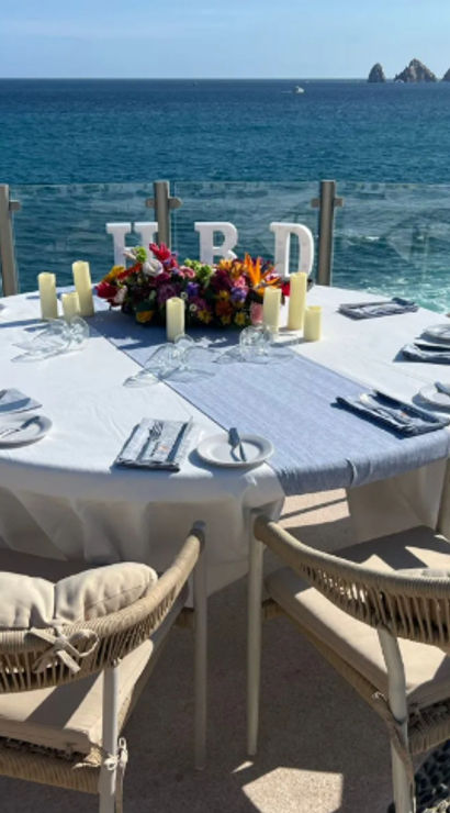 Sunlit oceanfront dining table set for six on a seaside terrace with cushioned wicker chairs, white linen, floral centerpiece and candles overlooking turquoise water and distant rocky headland.