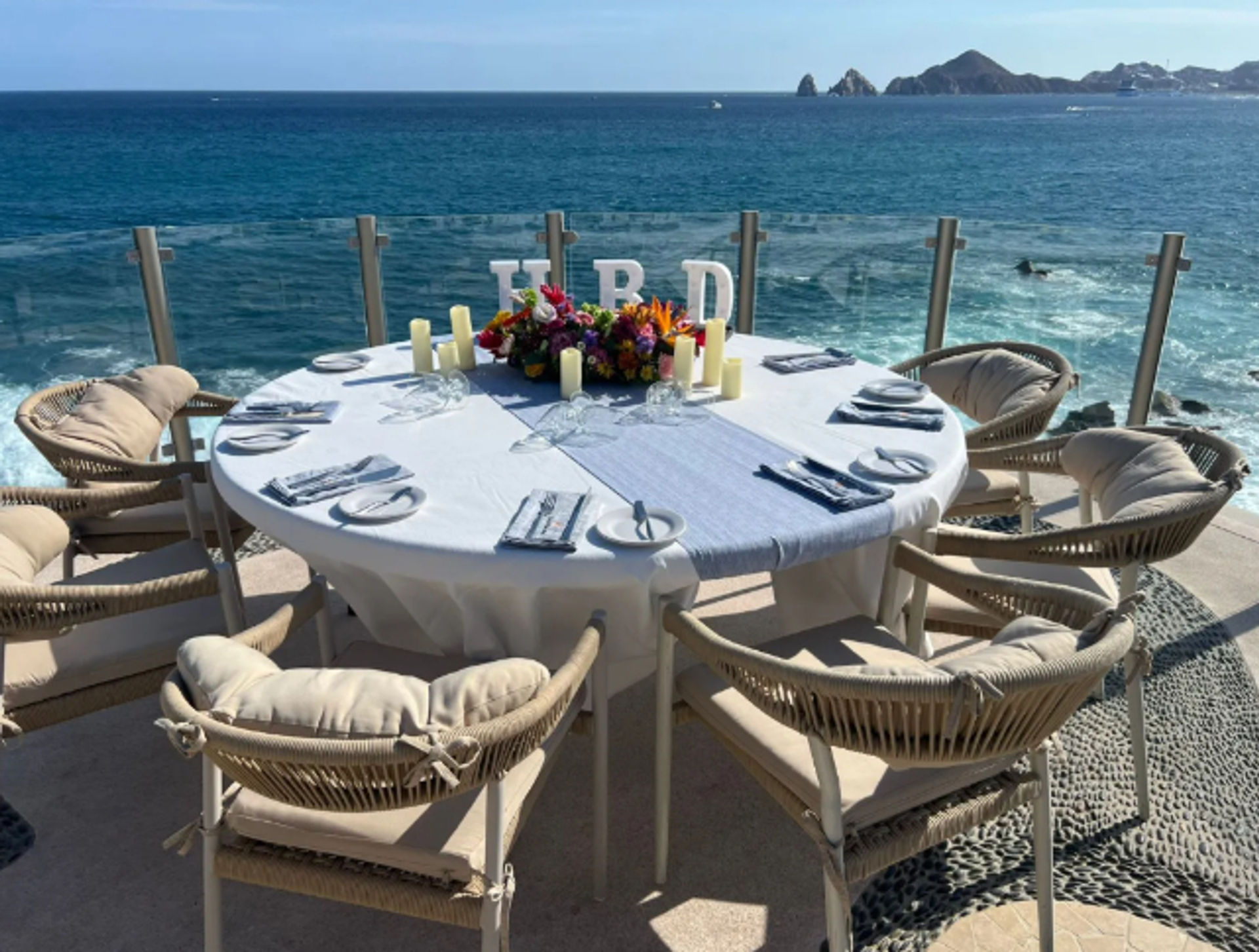 Sunlit oceanfront dining table set for six on a seaside terrace with cushioned wicker chairs, white linen, floral centerpiece and candles overlooking turquoise water and distant rocky headland.
