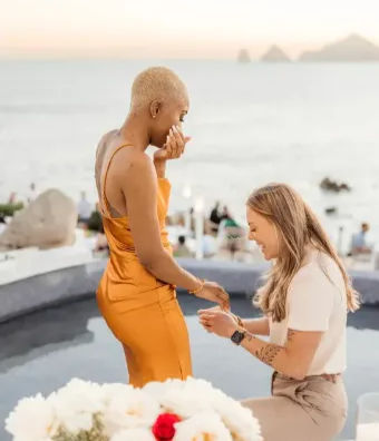 Romantic oceanfront sunset proposal — person kneeling to place a ring on their partner in an orange satin dress on a seaside terrace with white flowers in the foreground.