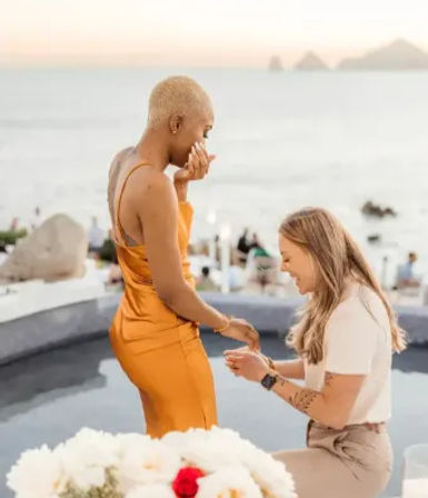 Romantic oceanfront sunset proposal — person kneeling to place a ring on their partner in an orange satin dress on a seaside terrace with white flowers in the foreground.