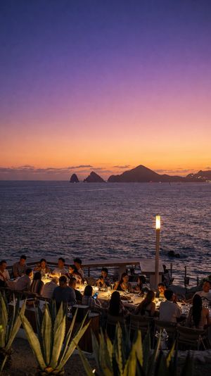 Oceanfront sunset dining scene—cozy candlelit tables by the water with friends, purple-orange sky and distant rocky islands on the horizon.