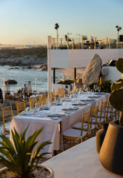 Oceanfront sunset dining on a beachside terrace with a long white-tablecloth table, gold chairs, and glassware overlooking a rocky coastline.