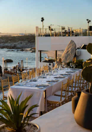 Oceanfront sunset dining on a beachside terrace with a long white-tablecloth table, gold chairs, and glassware overlooking a rocky coastline.