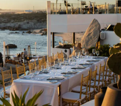 Oceanfront sunset dining on a beachside terrace with a long white-tablecloth table, gold chairs, and glassware overlooking a rocky coastline.