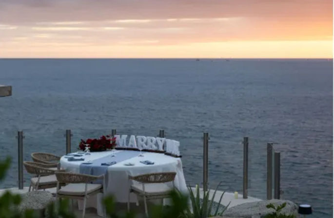 Romantic oceanfront sunset terrace with an intimate two-person table set for a proposal, white tablecloth, floral centerpiece and large 'MARRY' letters overlooking the calm sea.