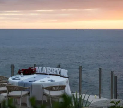 Oceanfront sunset proposal setup on a cliffside terrace: intimate table for two with white linens, bouquet, decorative 'MARRY' letters and glass railing overlooking the calm sea.