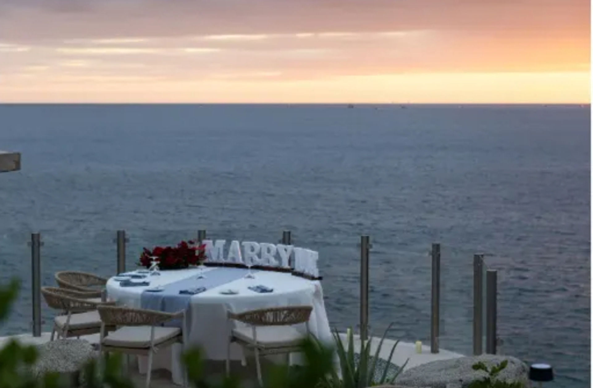 Oceanfront sunset proposal setup on a cliffside terrace: intimate table for two with white linens, bouquet, decorative 'MARRY' letters and glass railing overlooking the calm sea.