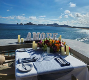 Romantic oceanfront table for two with AMORE letters, colorful floral centerpiece and candles on a seaside terrace overlooking blue water and distant rocky islands under a sunny sky