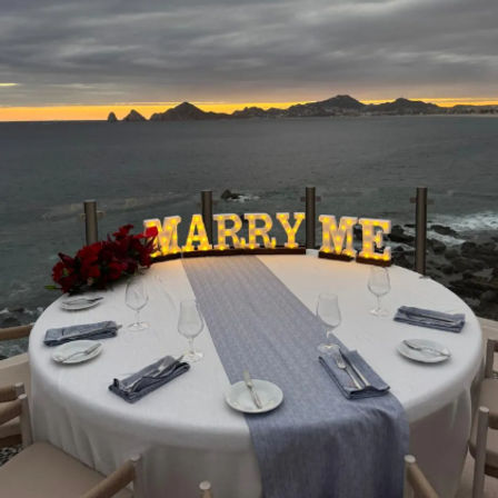 Romantic coastal proposal at sunset — round table for two on a cliffside terrace with illuminated “MARRY ME” letters, red roses, ocean view and rocky islets in the distance.