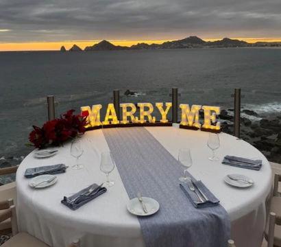 Romantic coastal proposal at sunset — round table for two on a cliffside terrace with illuminated “MARRY ME” letters, red roses, ocean view and rocky islets in the distance.