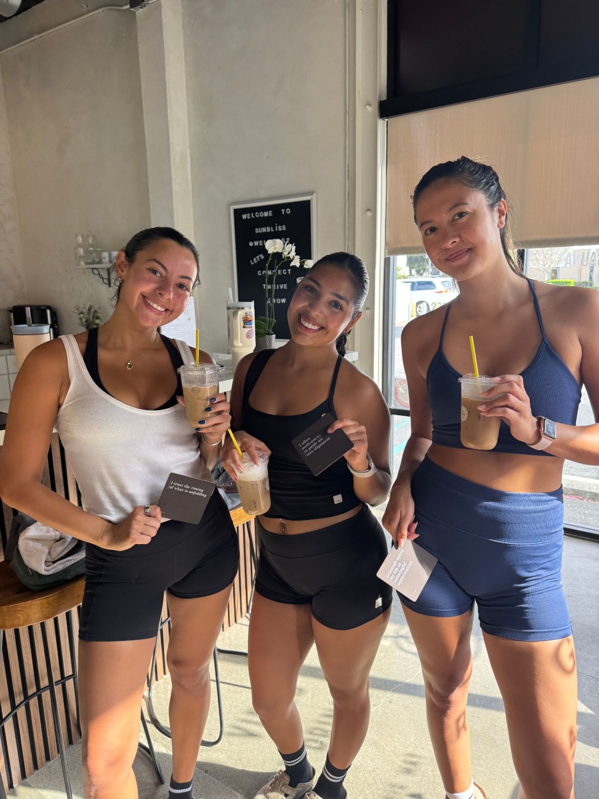 Three smiling friends in workout gear (sports bras and shorts) holding iced coffees and loyalty cards inside a sunlit cafe