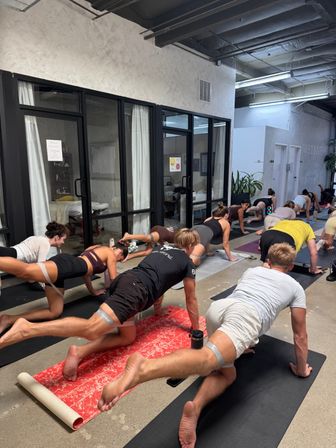 Group fitness class in a modern industrial-style wellness studio doing plank leg lifts on mats near glass-walled treatment rooms and potted plants.