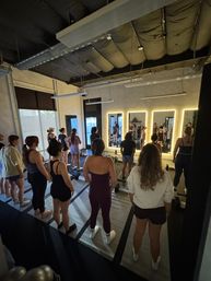 Participants standing on mats in an industrial-style urban fitness studio facing illuminated mirrors during a mat-based group workout class, warm lighting and high ceilings conveying an energetic vibe.