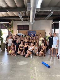 Large group of people in athletic wear posing and smiling in a bright industrial-style fitness studio with exposed ductwork, potted plants and a rolled blue yoga mat on the concrete floor — group fitness/yoga class photo.