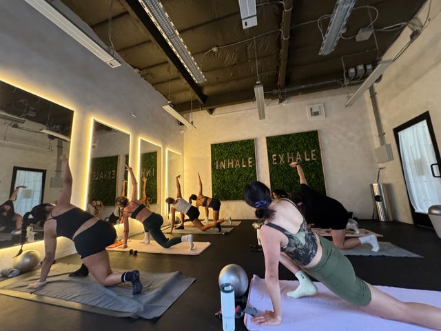 Group yoga class in a modern indoor fitness studio, participants stretching on mats in front of illuminated mirrors and green 'INHALE EXHALE' wall panels.