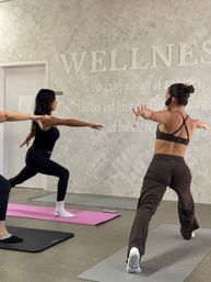 Group yoga class in a bright wellness studio — participants hold Warrior II poses on mats beneath a large 'WELLNESS' wall sign, instructor wearing a headset leads the session.