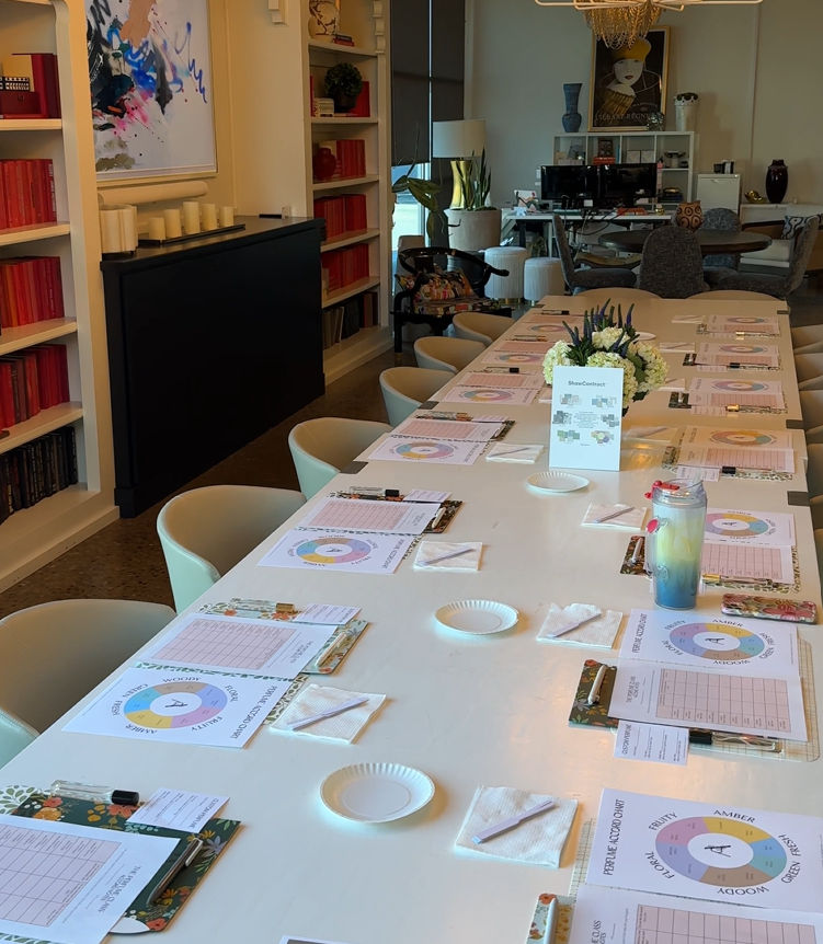 Long white conference table in a modern creative studio set for a workshop — clipboards, worksheets, pens, paper plates and floral centerpiece, pale chairs, bookshelves and artwork in background