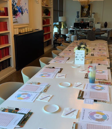 Long white conference table in a modern creative studio set for a workshop — clipboards, worksheets, pens, paper plates and floral centerpiece, pale chairs, bookshelves and artwork in background