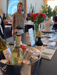 Ice bucket of wine bottles and canned drinks on a modern kitchen showroom island, with colorful flower bouquets, design samples on the counter, and blurred people chatting in the background.