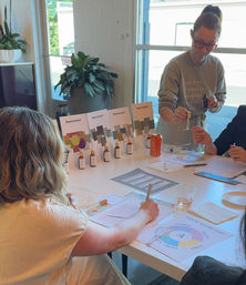 Sunlit design studio workshop with participants at a white table sampling small scent vials, filling worksheets and consulting color swatch brochures and a scent wheel.