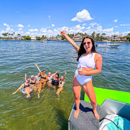 Group of friends in swimsuits partying from a motorboat into shallow coastal water, palm-lined waterfront homes and sunny blue sky