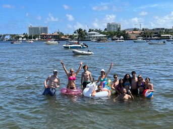 Group of people in swimsuits having fun in shallow coastal water on colorful inflatables (unicorn, pink donut, beach ball) with pleasure boats and waterfront homes on the distant shoreline under a sunny blue sky
