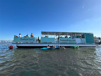 Turquoise pontoon party boat anchored in a sunny coastal bay, people socializing on deck and lounging on inflatable floats in clear blue water and sky.