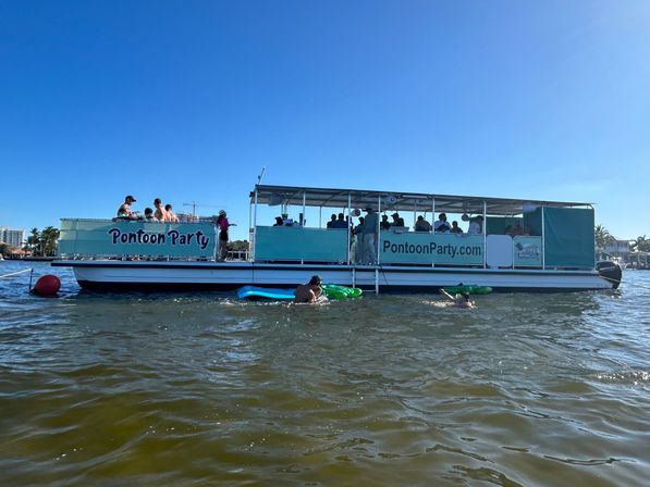 Teal party pontoon boat with people on board and swimmers on inflatables under a clear blue sky in a sunny coastal inlet near waterfront homes.