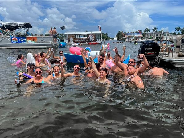 Cheering group of friends partying in shallow coastal water near anchored boats and pontoons, raising drinks and colorful inflatables under a sunny palm-lined waterfront.